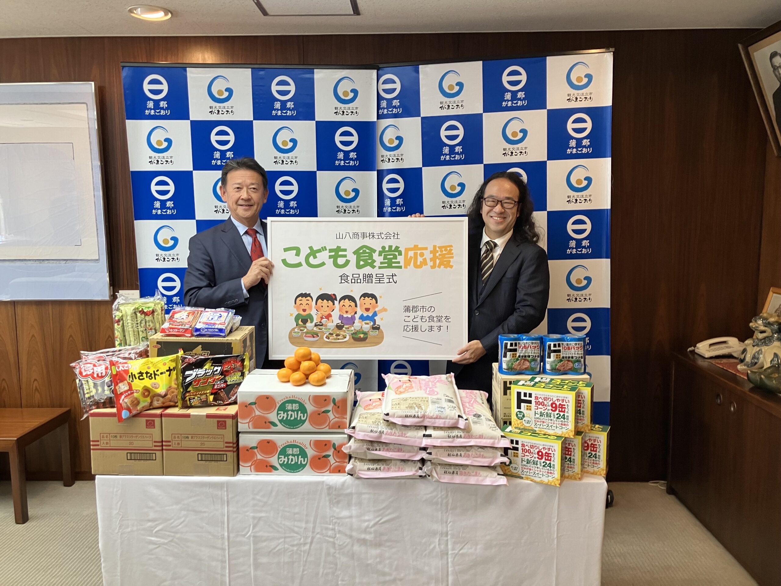 鈴木蒲郡市長(左)と鈴木弊社代表取締役(右)による贈呈式の様子。蒲郡みかんやお米など、多くの食品が並びました。